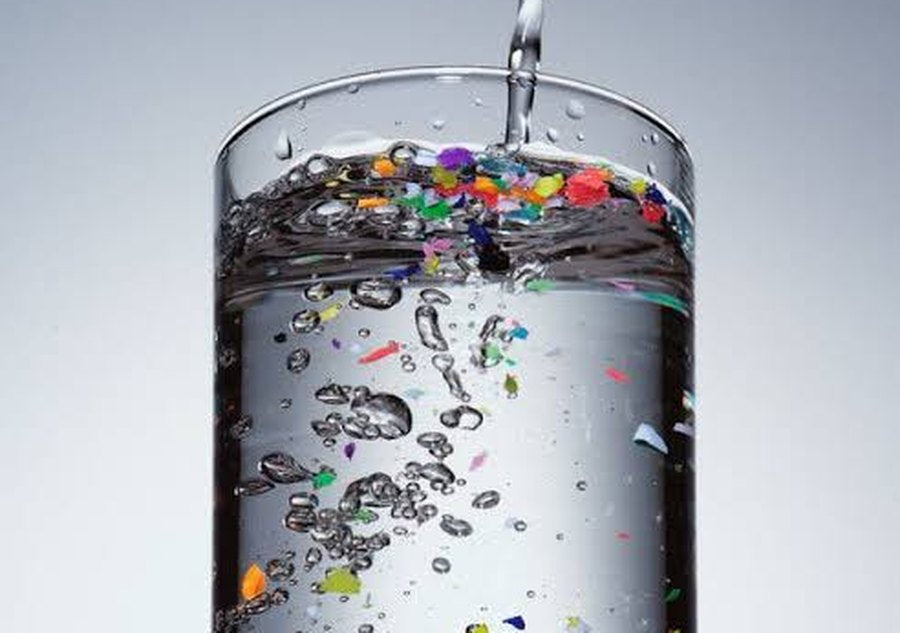 A tall glass of water with colourful microplastic particles floating in it, one cluster magnified in a zoom lens
